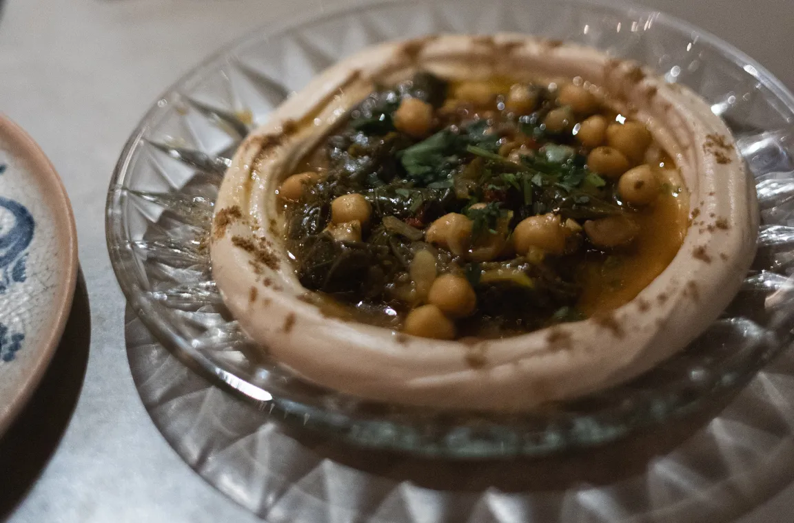Glass plate of hummus topped with chickpeas, greens, herbs, and olive oil, served as a mezze appetizer.