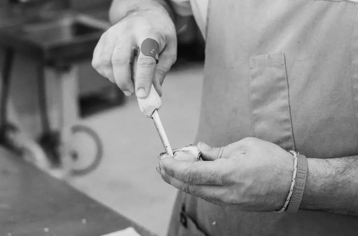 Black-and-white close-up of a chef shucking a fresh oyster by hand, wearing an oyster knife glove and apron at Little Em's Oyster Bar.