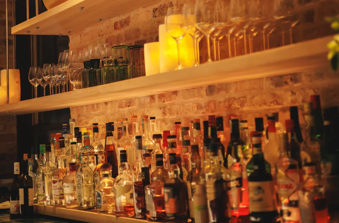 Warmly lit bar shelves displaying an assortment of spirits, wine glasses, and candles against an exposed brick wall.