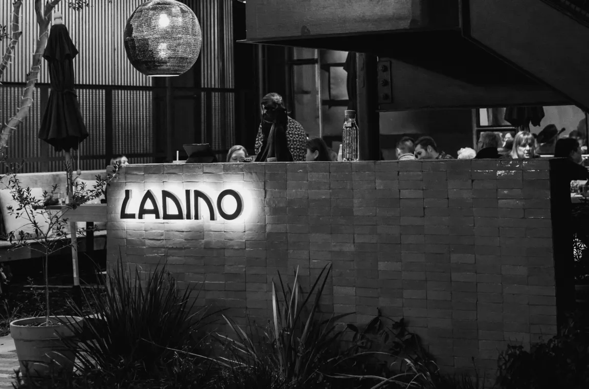 Black-and-white evening view of Ladino restaurant patio with glowing sign and diners gathered outdoors.