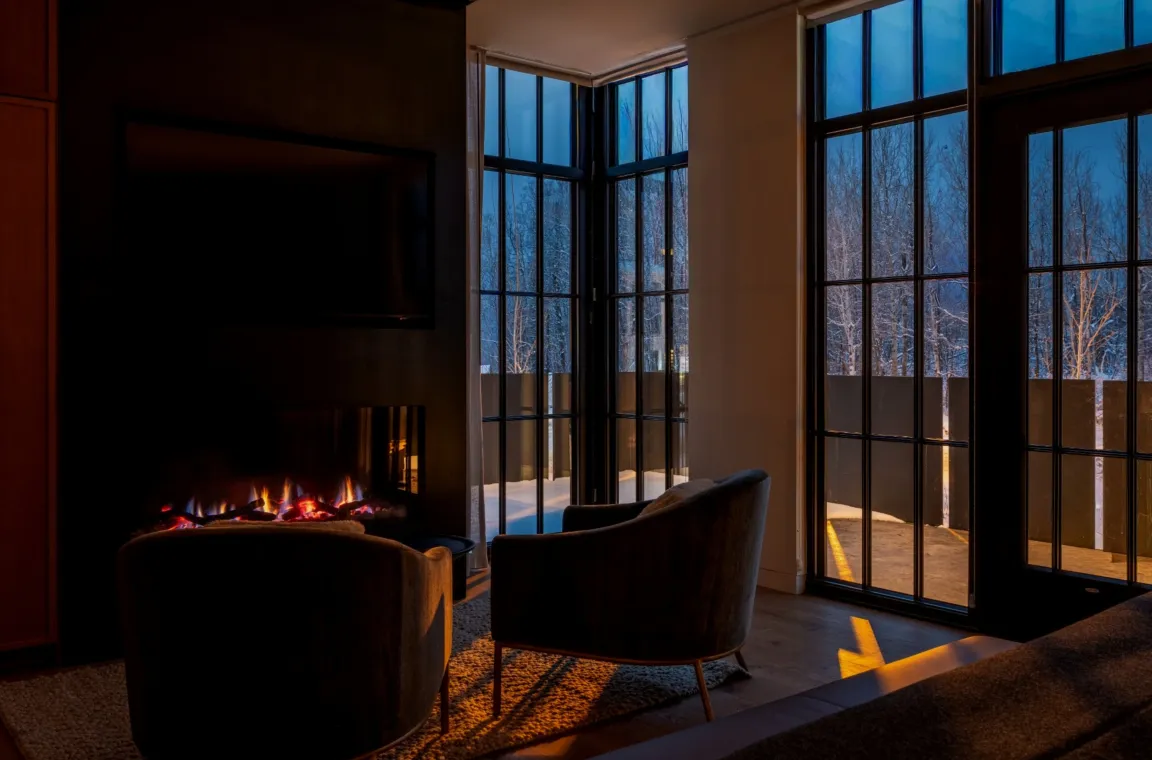 Floor-to-ceiling windows frame a snowy forest beyond a glowing fireplace inside a Larch House guest suite.