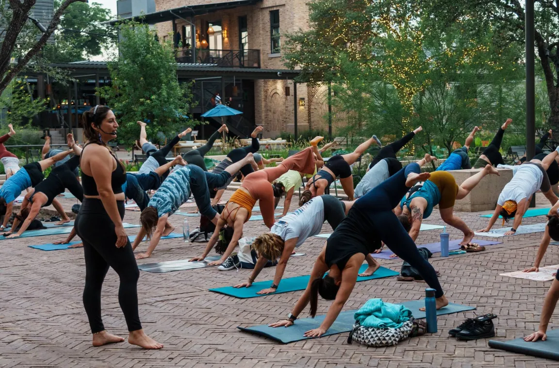 Outdoor community yoga class at Pearl in San Antonio, with participants practicing on mats in the plaza beneath trees wrapped in twinkling lights.