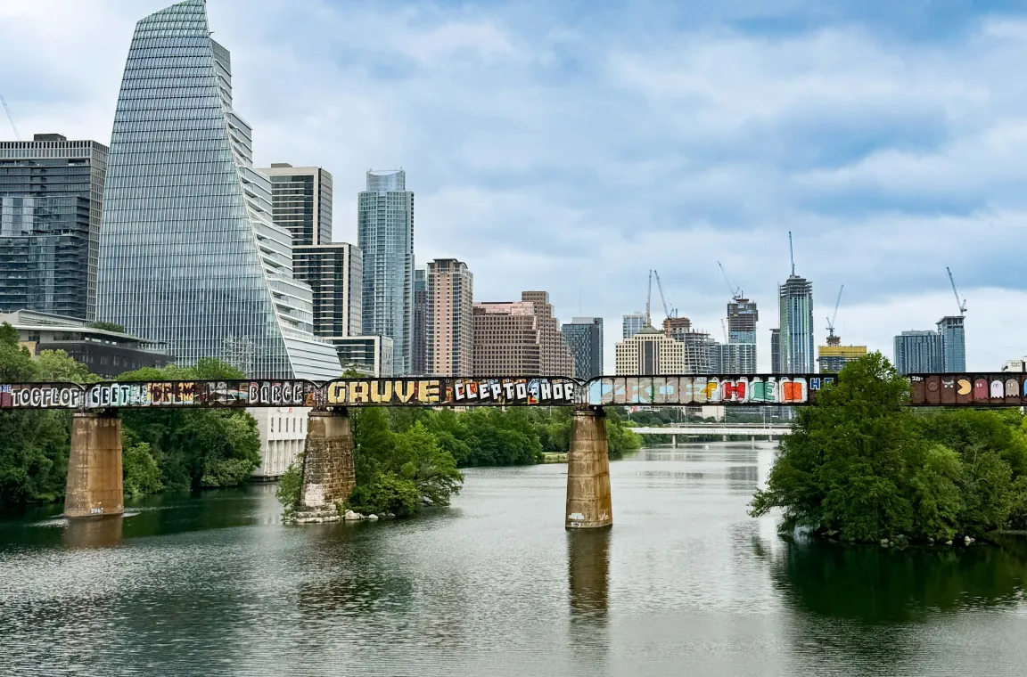 View of Lady Bird Lake with the Austin skyline and a graffiti-covered railroad bridge spanning the river beneath a cloudy sky.