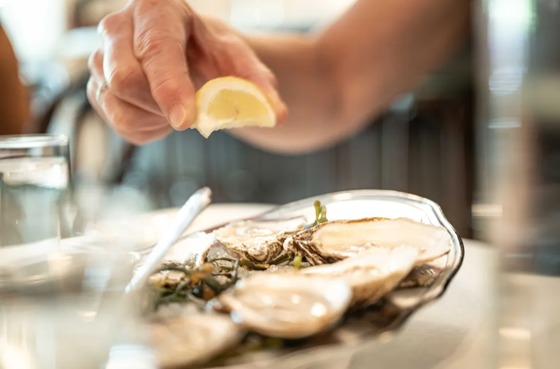 Hand squeezing a fresh lemon wedge over a platter of raw oysters served on ice with seaweed garnish.