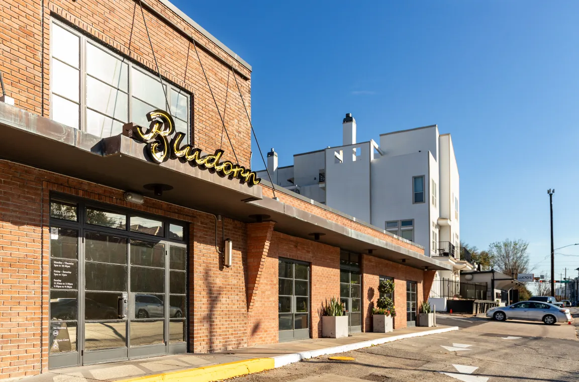 Exterior view of Bludorn restaurant in Houston, featuring a historic brick facade, large glass doors, and a glowing gold script Bludorn sign under a clear blue sky.