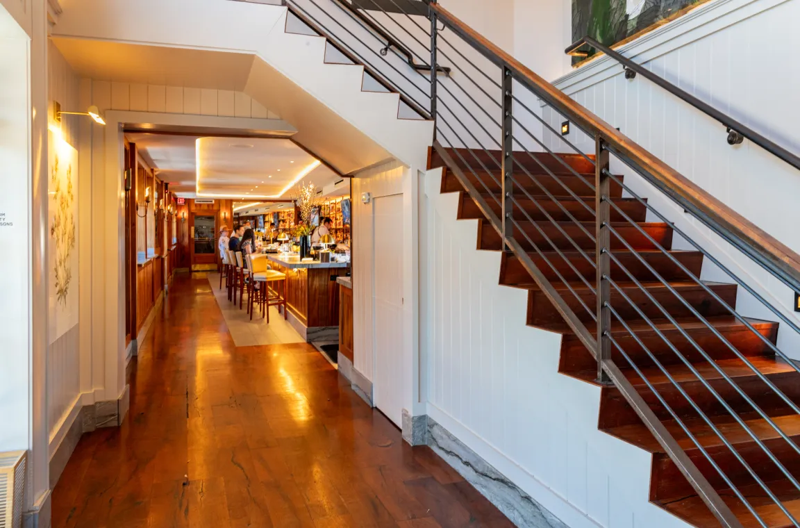 Interior of Bludorn restaurant showing a rich wood staircase with a metal railing next to a long hallway leading to a dimly lit bar area with patrons.
