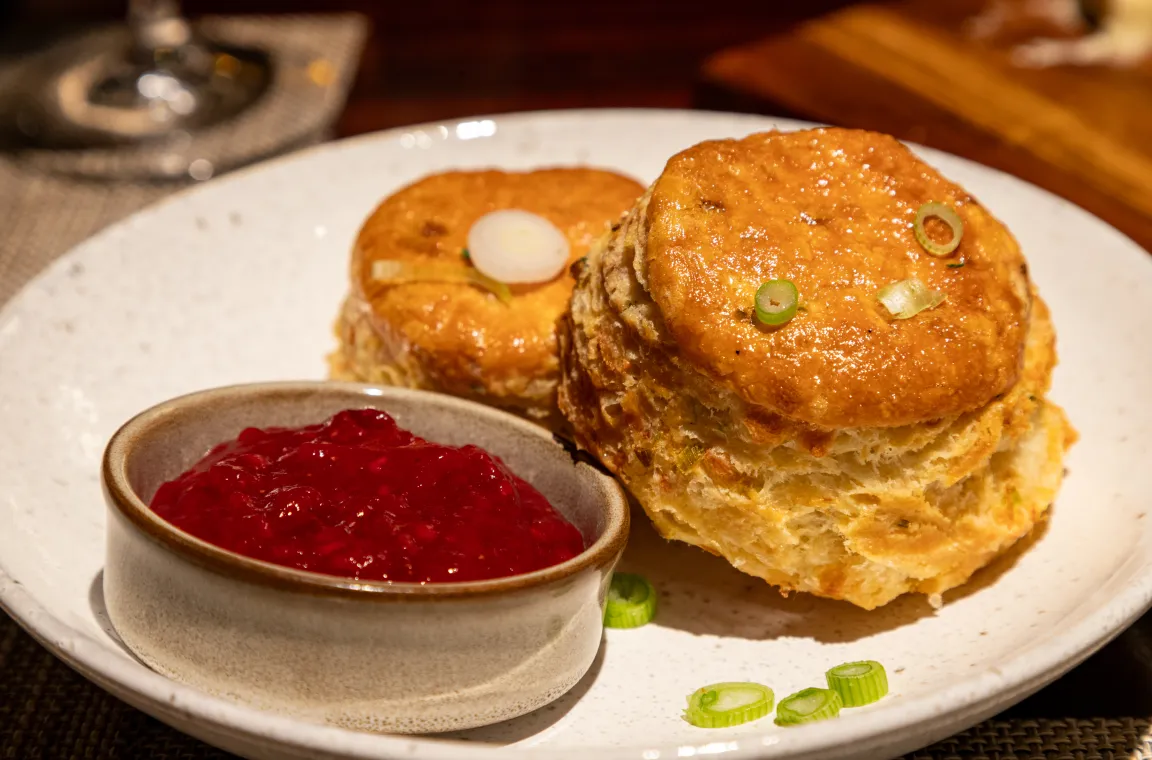 Close-up of two golden-brown, flaky biscuits garnished with sliced green onions on a white plate, served with a small bowl of vibrant red raspberry jam.