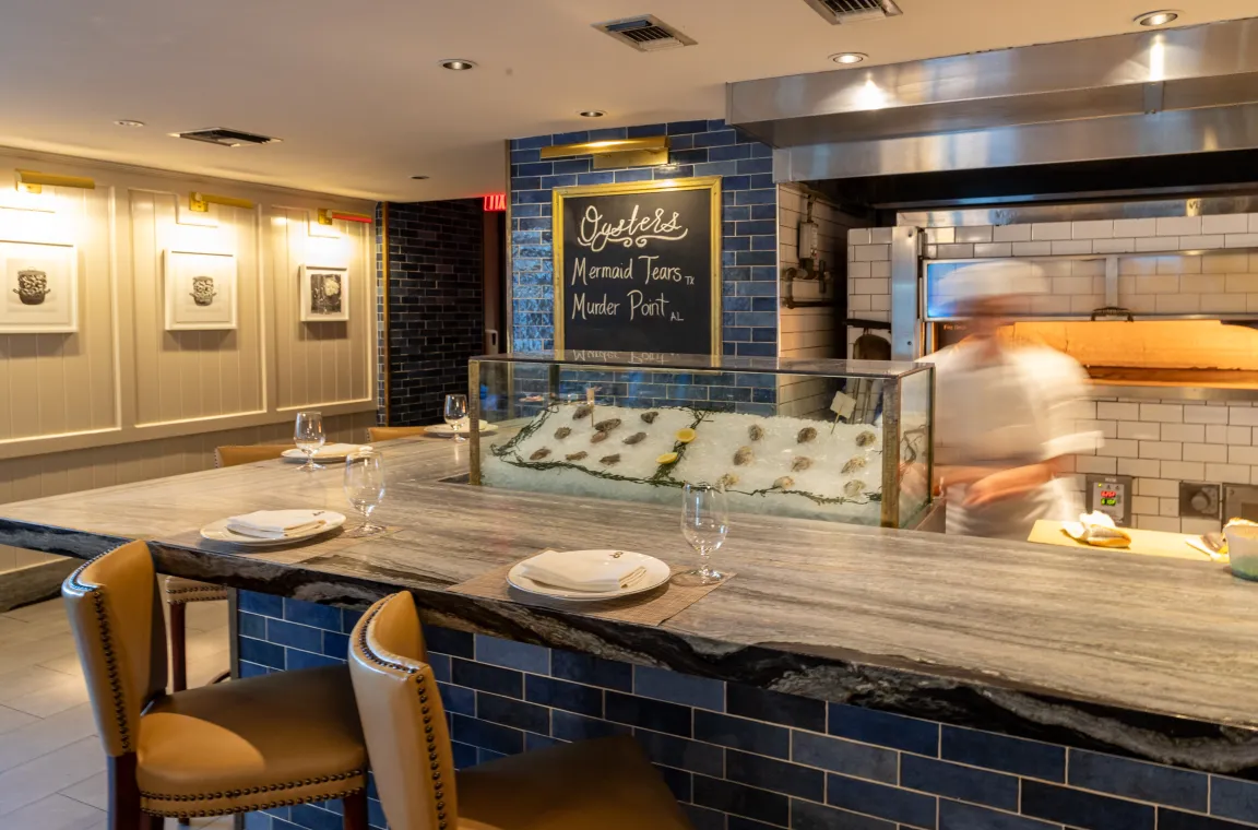 The oyster bar at Bludorn, featuring an iced display of fresh oysters, a chalkboard listing Mermaid Tears and Murder Point varieties, and a chef working near a wood-fired oven.