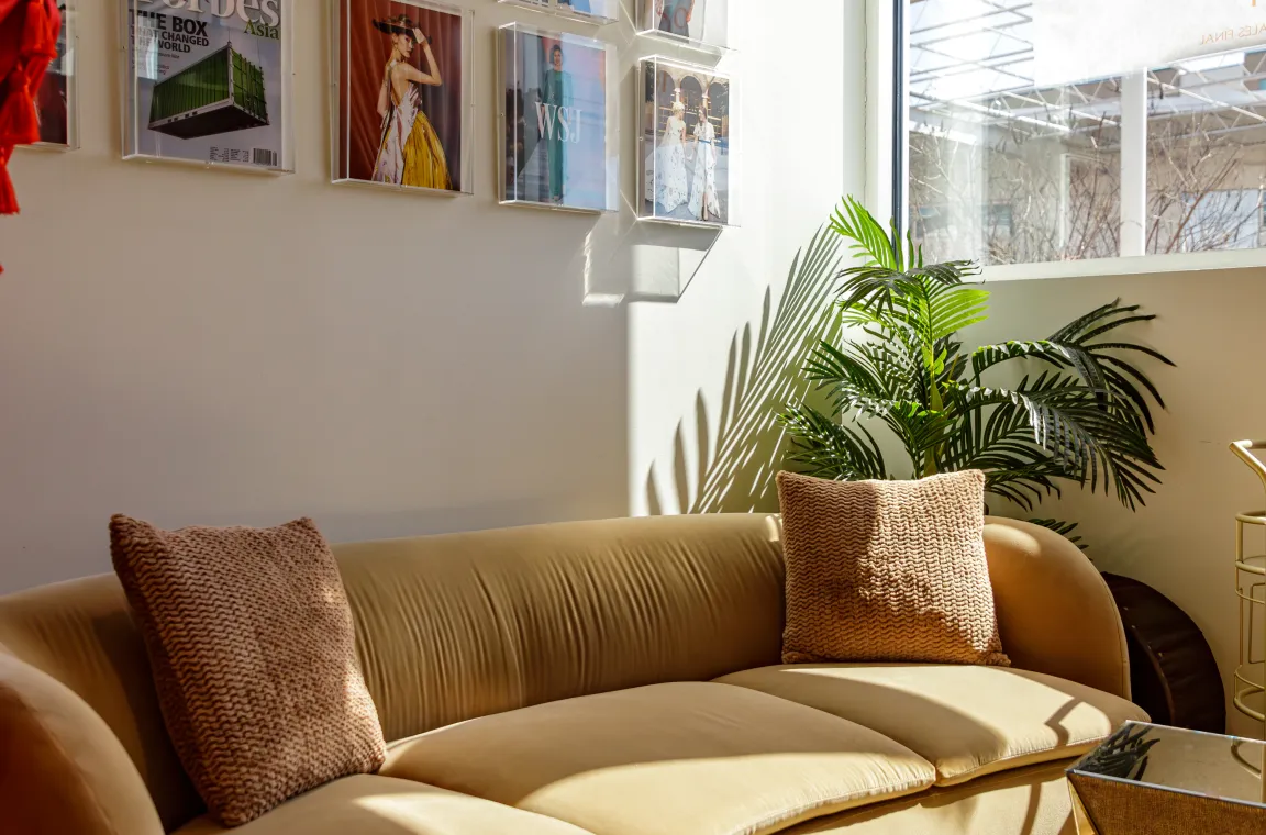 Inside the Chloe Dao boutique, featuring a plush tan velvet sofa with brown throw pillows illuminated by sunlight, set against a wall decorated with framed magazine covers.