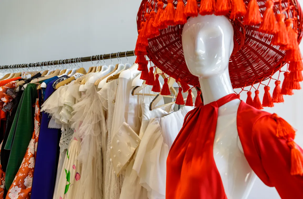 A close-up of a white mannequin wearing a vibrant red dress and a matching oversized straw hat with tassels, standing in front of a clothing rack filled with designer garments.