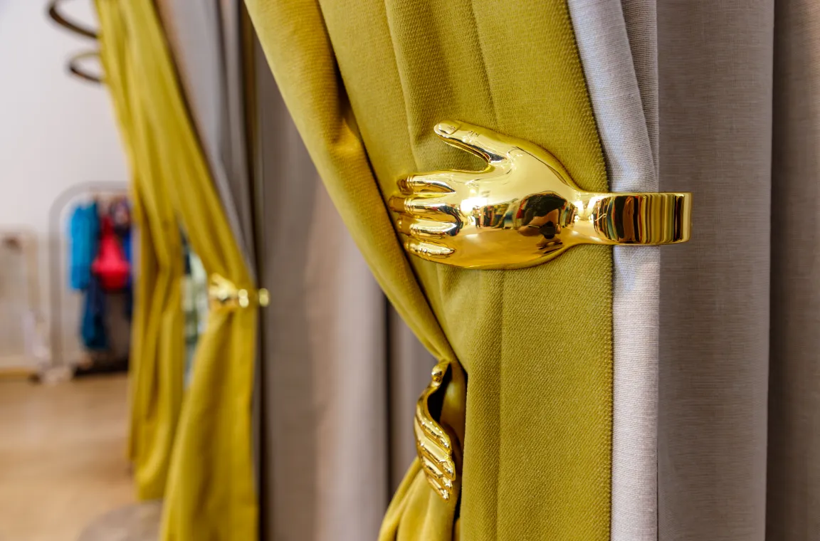 A detailed close-up of heavy mustard-yellow and beige dressing room curtains held open by a unique, polished gold bracket shaped like a human hand.