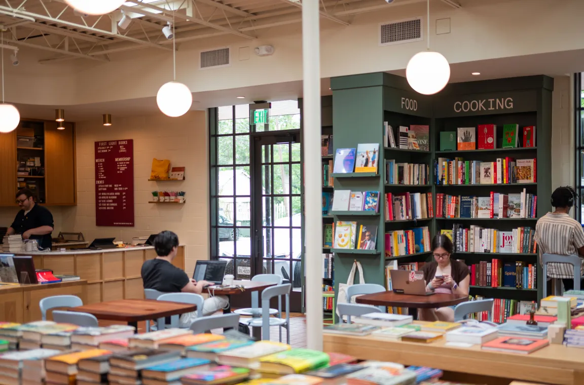 Interior of First Light Books featuring curated bookshelves, reading tables, a checkout counter, and customers browsing, reading, and working in a bright, welcoming space.
