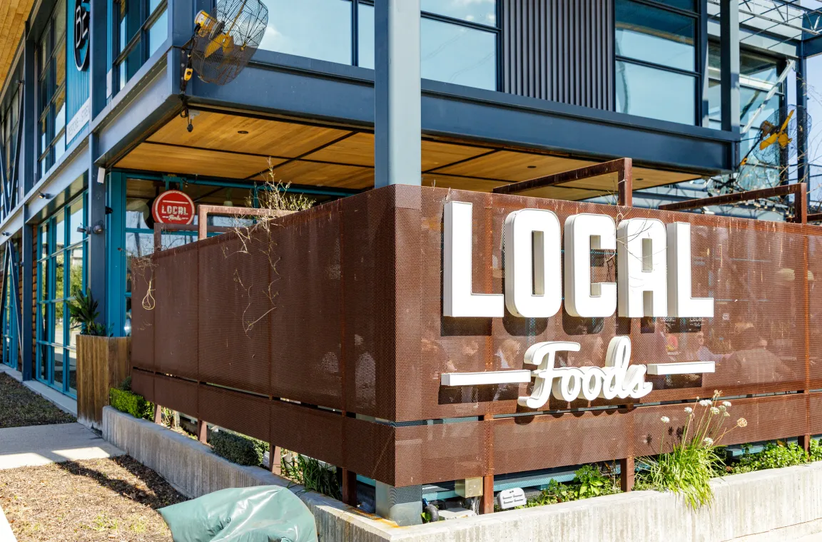 Exterior storefront of Local Foods along the Heights Hike and Bike Trail, featuring a prominent logo on a rusted metal mesh patio wrapper.