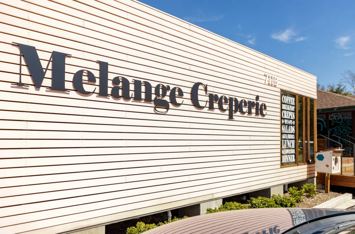 Large black text signage for Melange Creperie mounted on the light-colored horizontal wood siding of its storefront.