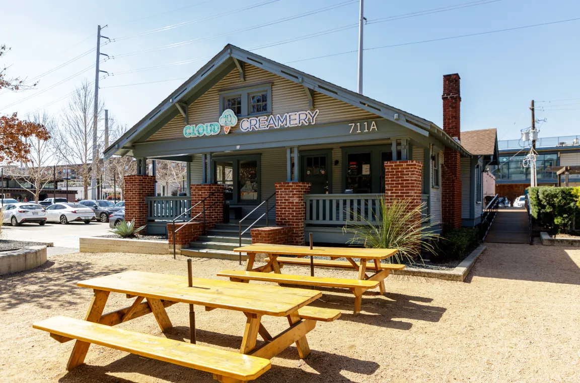 A charming, light blue bungalow style building housing Cloud 10 Creamery, featuring brick pillars, a front porch, and wooden picnic tables in a gravel courtyard.
