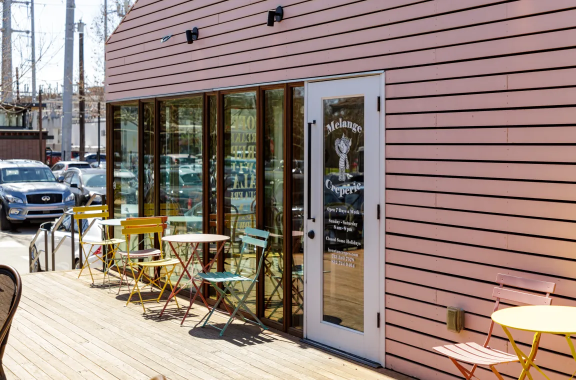 The outdoor wooden deck and glass entrance of Melange Creperie, featuring colorful metal bistro chairs and tables set against pink horizontal siding.