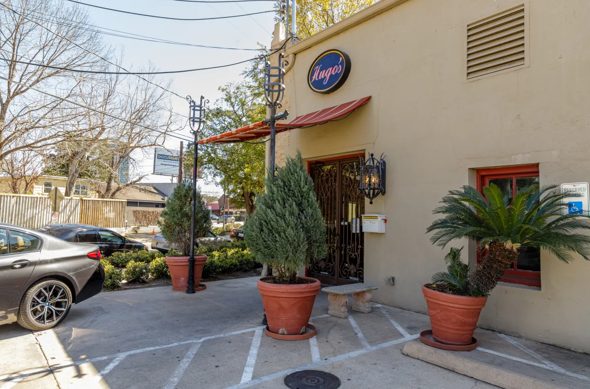 Exterior view of Hugo's restaurant in Houston, featuring a tan stucco building with arched iron doors, a striped awning, and large potted plants at the entrance.
