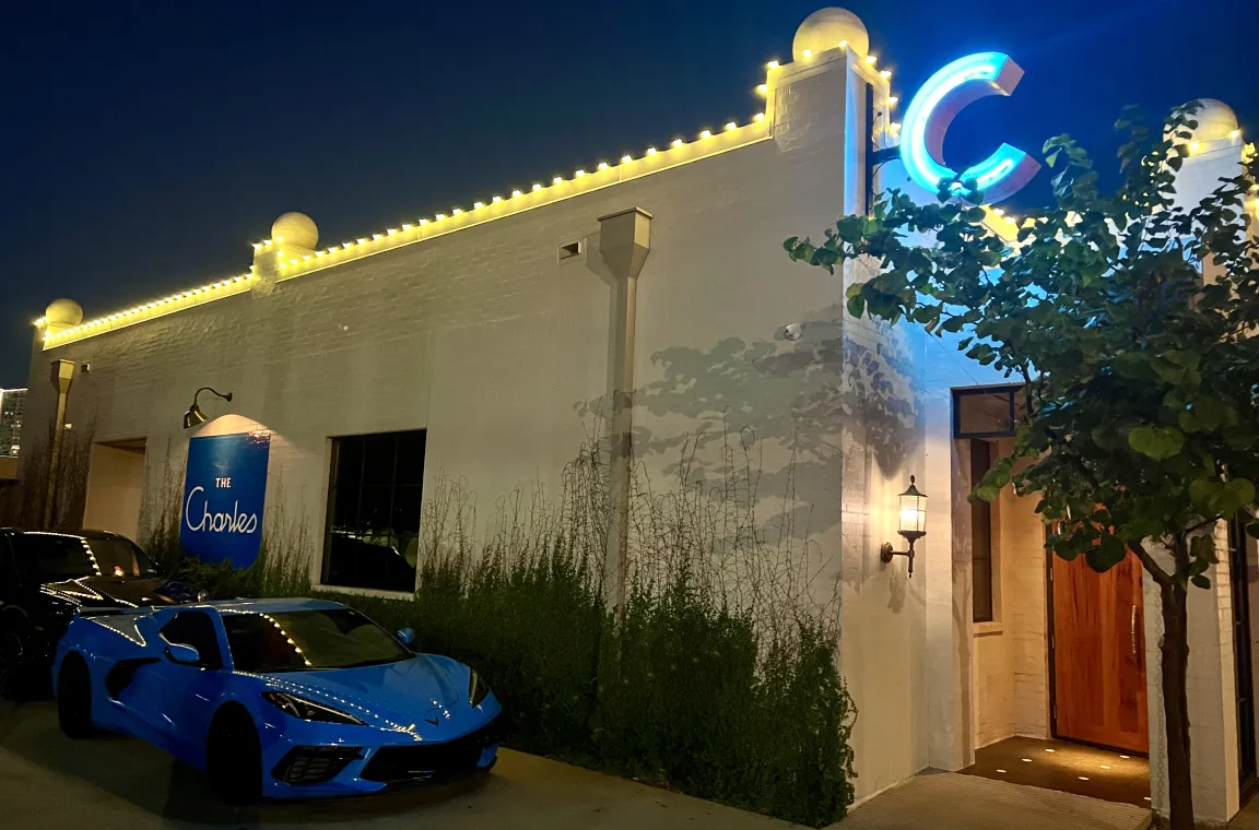 The Charles restaurant illuminated at night with glowing roofline lights and neon signage, as a blue sports car is parked outside the entrance.