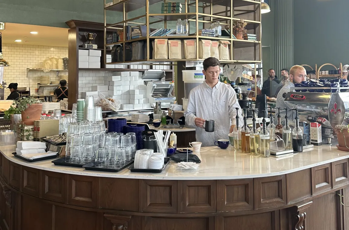A busy bakery cafe interior featuring a curved, dark wood-paneled coffee bar with a white marble countertop, where a barista works underneath a large hanging gold geometric shelf unit filled with coffee bags and books.