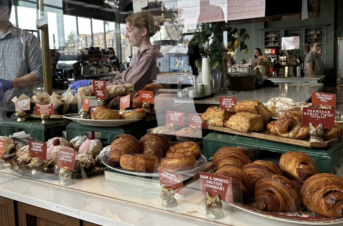 A vibrant glass display case inside a bakery filled with rows of freshly baked pastries, including croissants, morning buns, and scones, marked with red labels.
