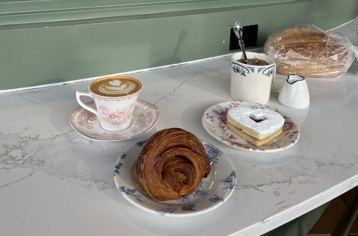 A scenic counter setup next to a bakery window showcasing a latte in a pink patterned teacup, a golden brown croissant, a heart-shaped cookie, and a fresh loaf of bread on vintage-style floral plates.