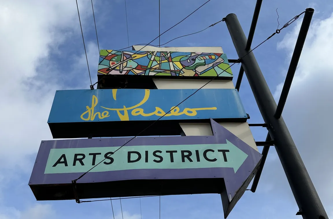 A retro, multi-tiered street sign mounted on a black pole against a cloudy blue sky, featuring a colorful mosaic pattern at the top, a blue section reading "the Paseo," and a purple arrow pointing right that reads "ARTS DISTRICT."
