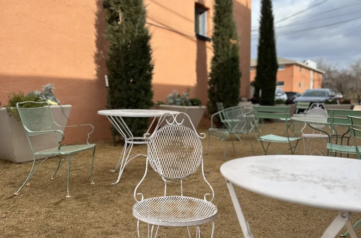 An outdoor gravel courtyard patio featuring white and light green metal filigree tables and chairs set against a tall peach-colored building with slender cypress trees.