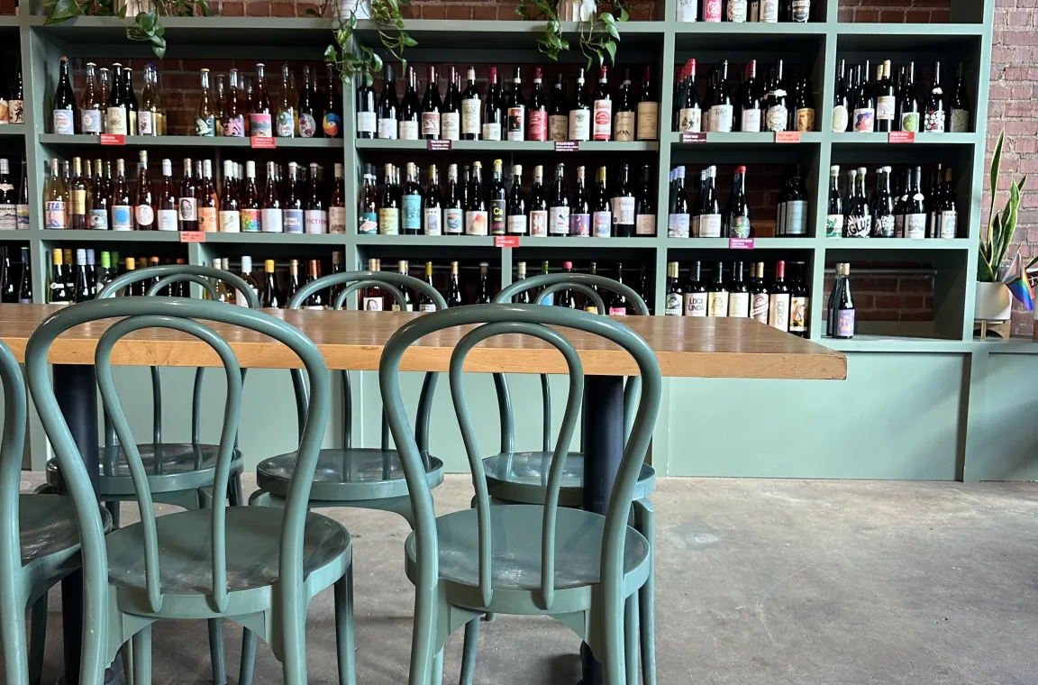 A wooden dining table surrounded by sage green bentwood chairs, situated in front of a large, matching sage green wall shelf stacked with rows of wine bottles against an exposed brick wall.