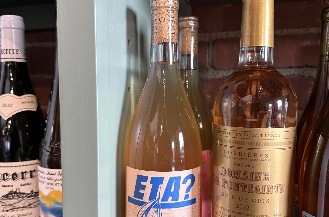 A close-up shot of wine bottles on a light green shelf, prominently displaying a bottle of rosé with a blue and pink label featuring a sailboat and the text "ETA? ROSÉ 2022."