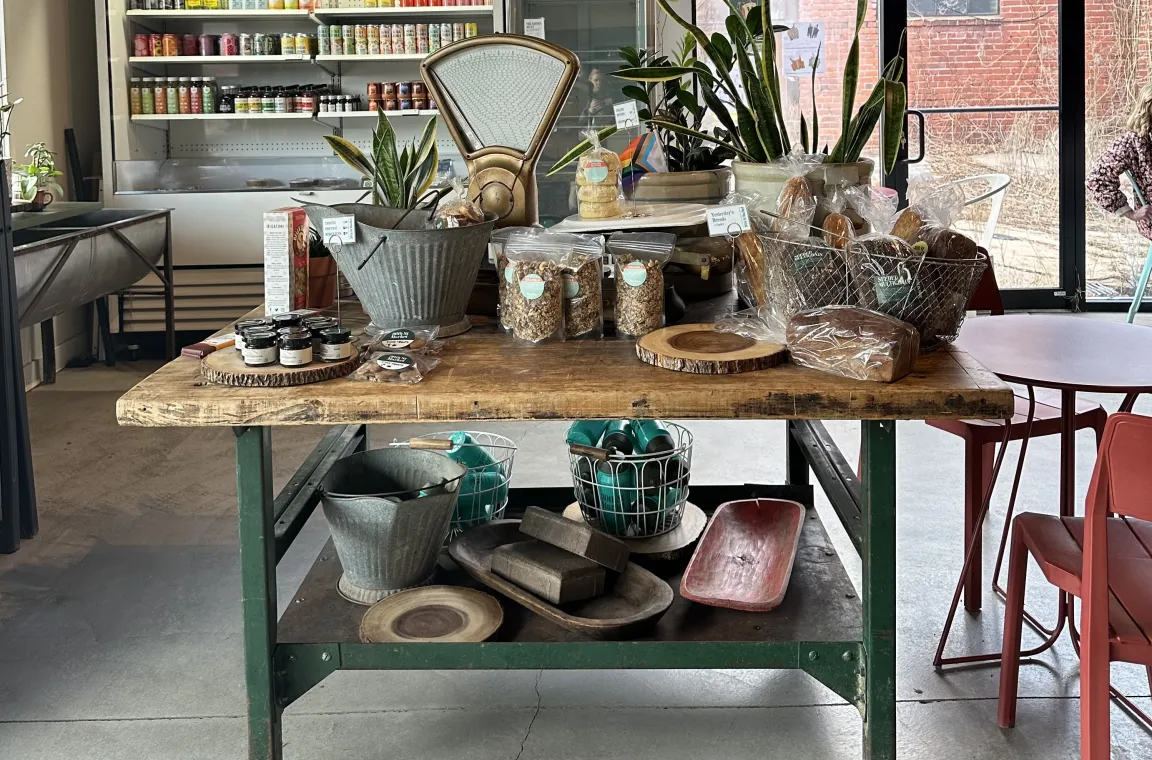 A rustic wooden display table inside a market, filled with packaged snacks, baked goods, and a vintage scale, positioned in front of "GRAB & GO" refrigerated coolers and a glass door.