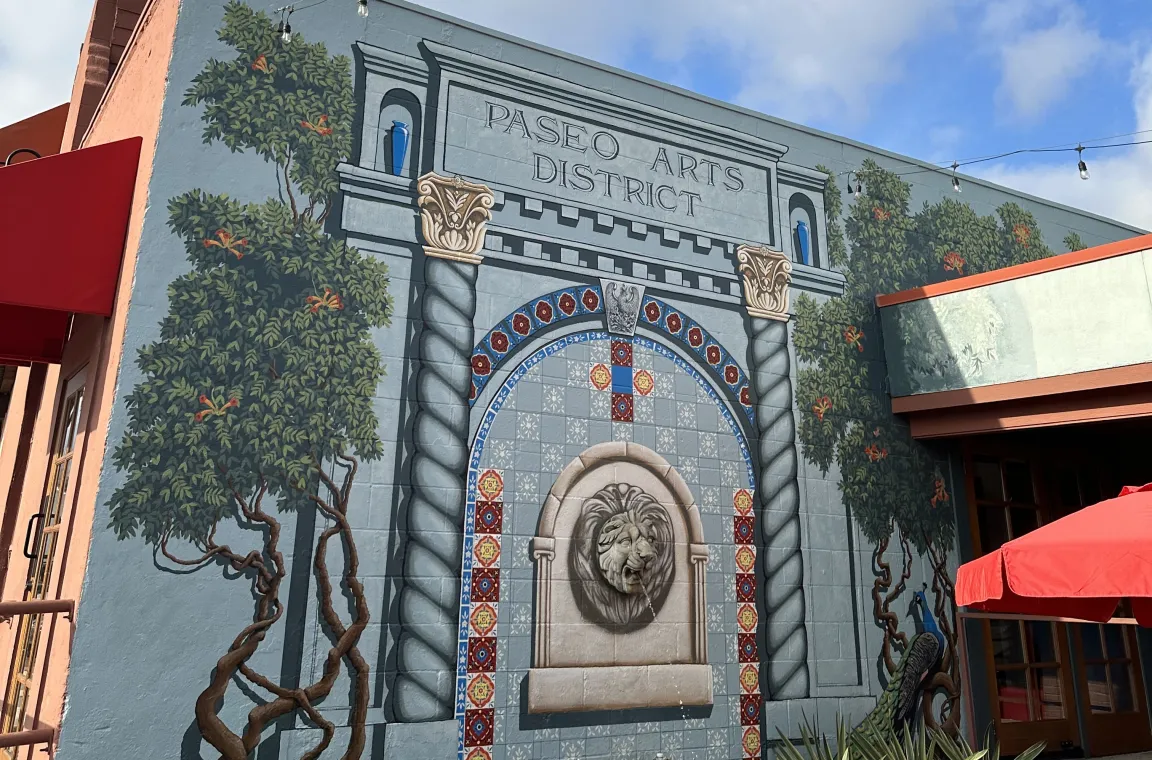 A large, detailed building mural depicting a classical stone archway with a lion's head fountain, flanked by painted trees, a peacock, and the text "PASEO ARTS DISTRICT" at the top.