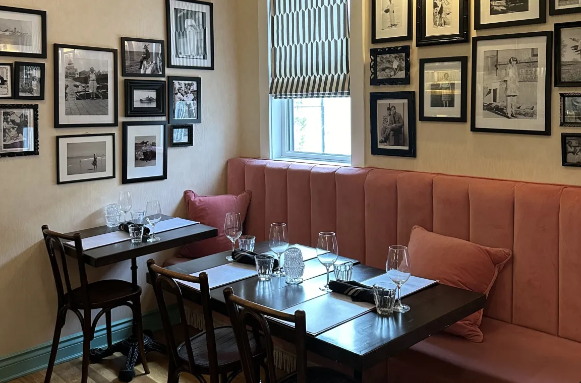 A cozy dining corner featuring two dark wood tables set with wine glasses, backed by a long, channeled dusty pink banquette and walls adorned with numerous framed black-and-white photographs.