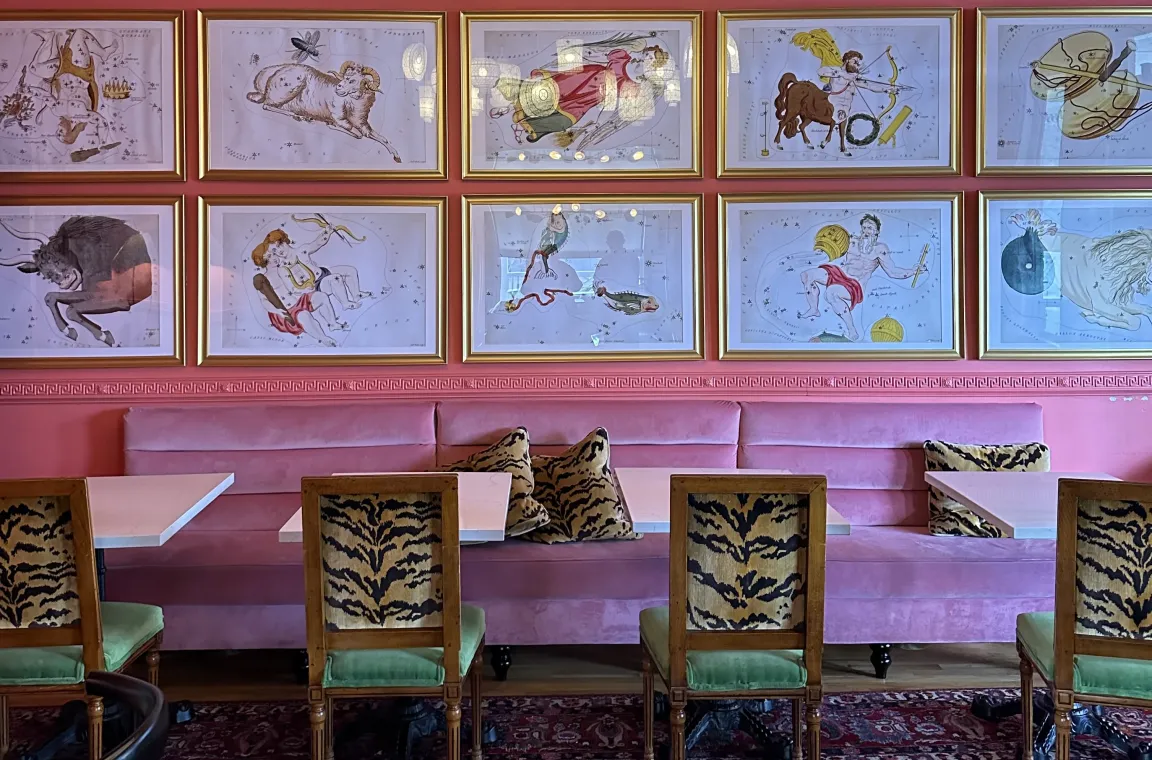A vibrant dining space with a long pink velvet banquette against a coral-colored wall displaying ten gold-framed illustrations of astrological constellations, paired with wooden chairs upholstered in tiger-print fabric and green velvet seats.