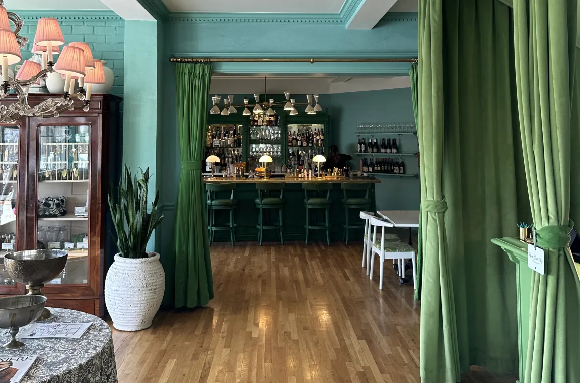 An elegant bar area viewed through parted green velvet curtains, showcasing a rich teal and seafoam green color scheme, a dark wood-topped bar with matching teal stools, hardwood floors, and a snake plant in a textured white pot.