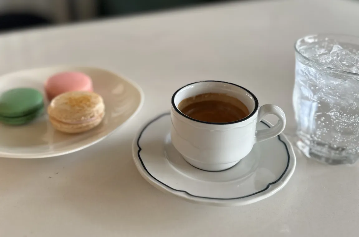 A close-up shot of a small white espresso cup and saucer with a black rim on a table, sitting next to a glass of iced water and a white plate holding three colorful macarons (green, pink, and tan).