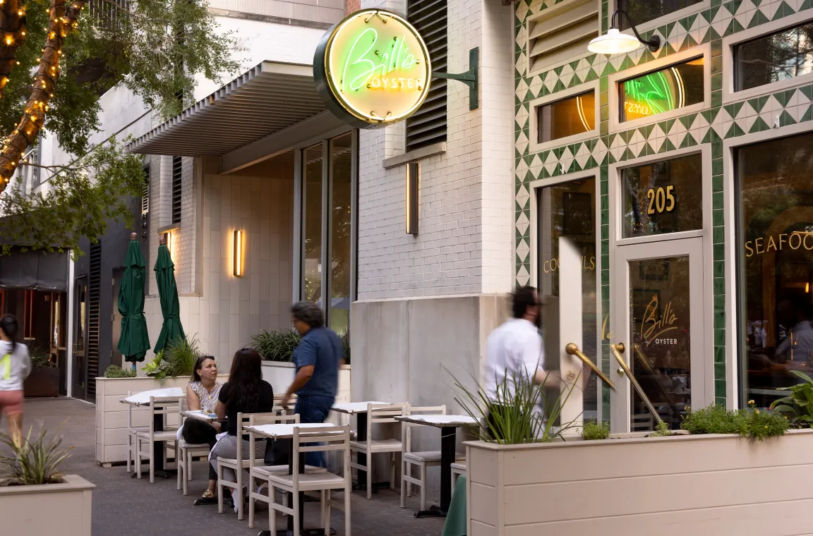 A restaurant storefront featuring outdoor seating, people seated at tables, and a waiter entering the business