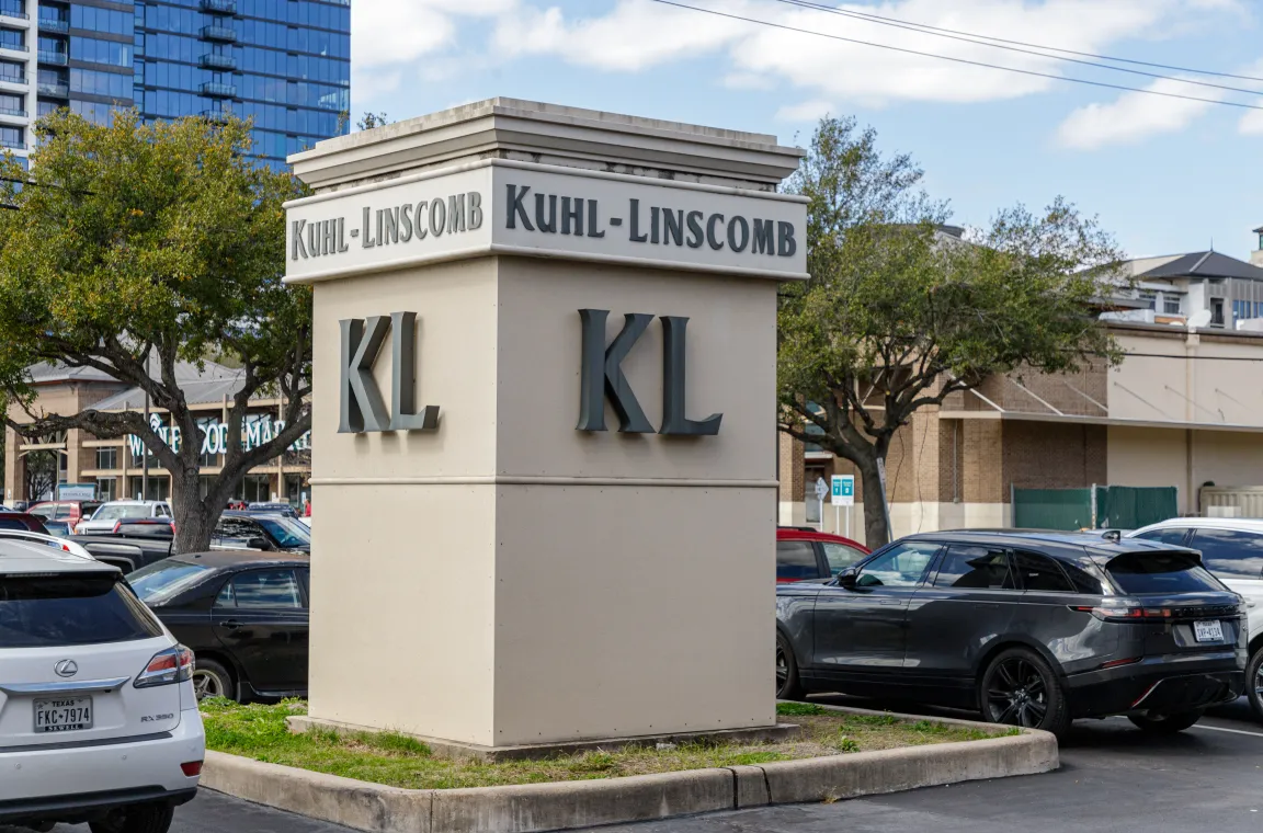A large beige monument sign with the letters KL and name Kuhl-Linscomb standing in a parking lot with cars and a Whole Foods Market in the background.