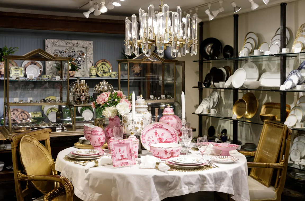 A formal dining room display featuring a table set with pink and white patterned chinaware, crystal wine glasses, and a crystal chandelier hanging overhead.