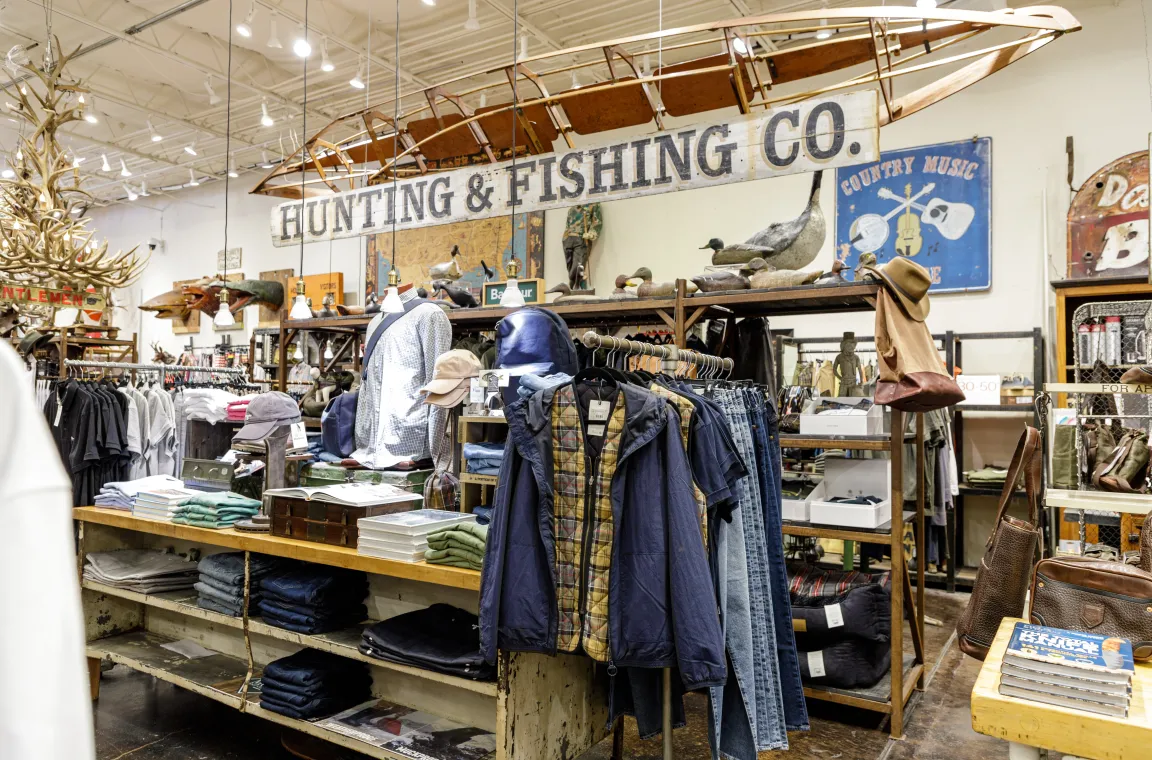 An upscale apparel display section with a rustic "Hunting & Fishing Co." sign hanging below an old wooden canoe, surrounded by jackets, jeans, and outdoor accessories.