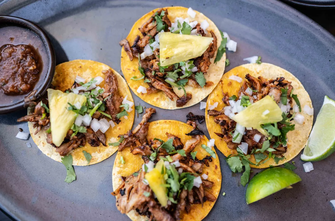 Four street-style tacos topped with grilled meat, pineapple, diced onion, and cilantro, served with salsa and fresh lime wedges at La Popular.