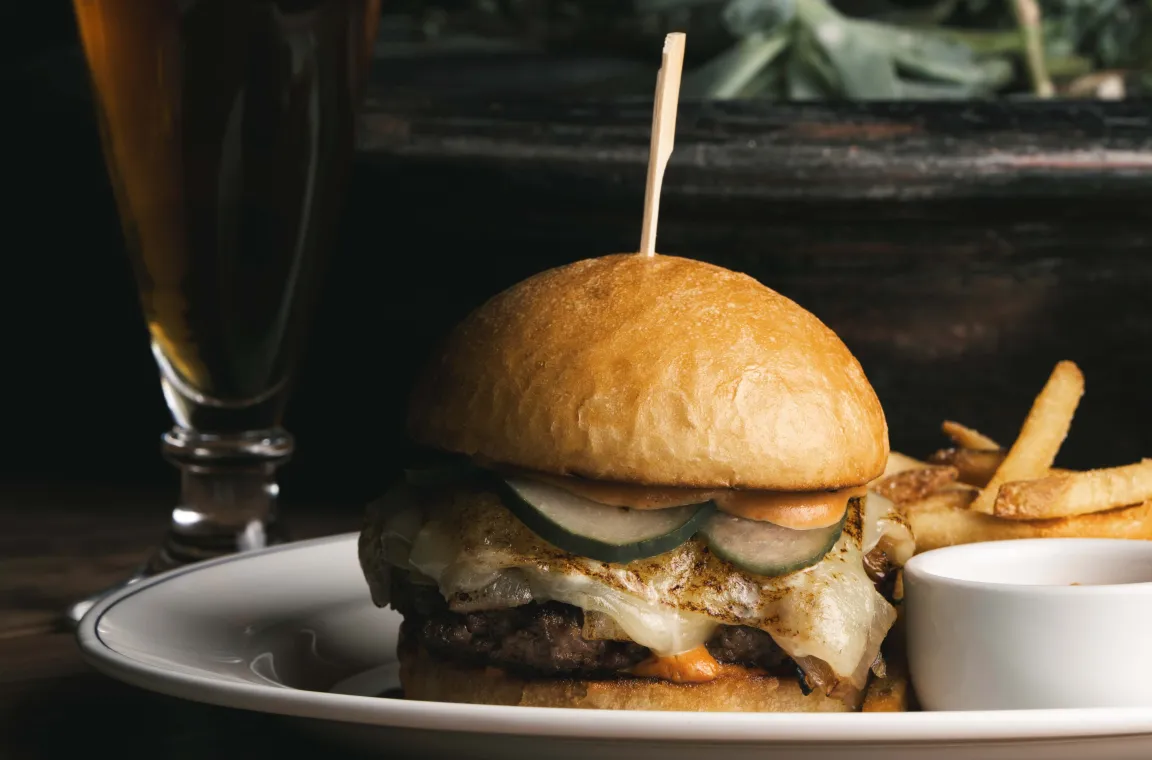 Gourmet cheeseburger with melted cheese, pickles, fries, and dipping sauce served alongside a dark craft beer.