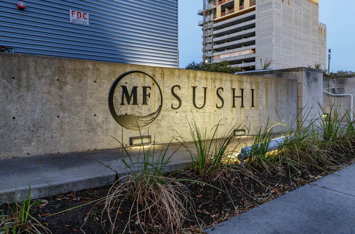 A concrete monument sign embedded with the black "MF SUSHI" circular logo and text, lit from below by landscape lighting in front of a modern building facade.