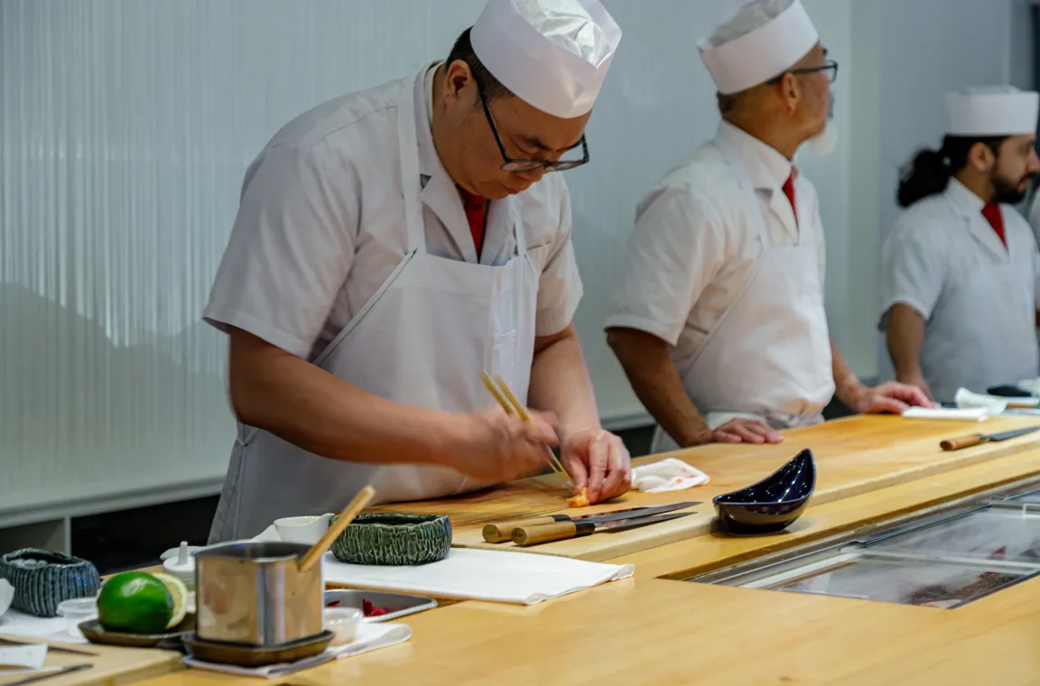 A sushi chef in a white uniform and traditional cap carefully assembling a dish with chopsticks behind a long wooden sushi counter alongside other chefs.