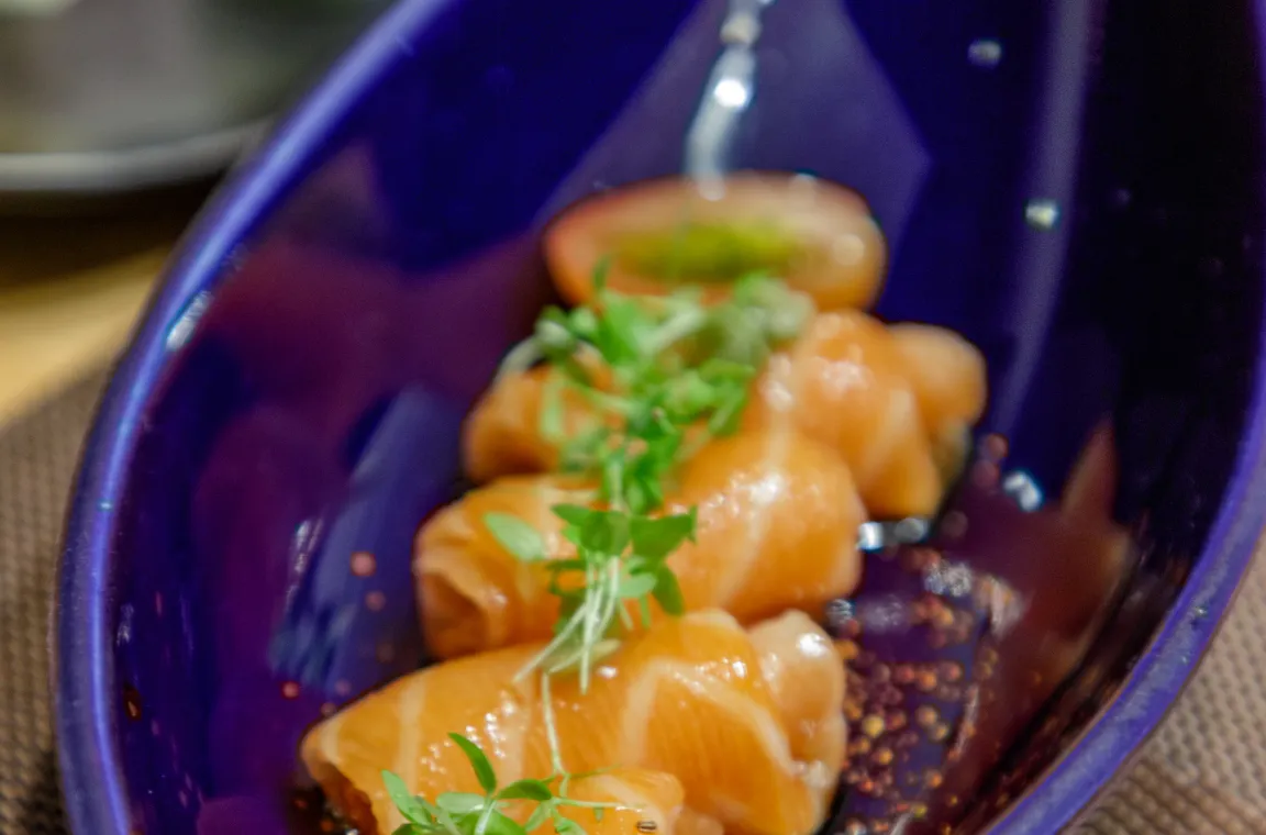 A close-up shot of raw salmon slices rolled up and served in a deep blue, boat-shaped dish garnished with fresh microgreens and cherry tomatoes.