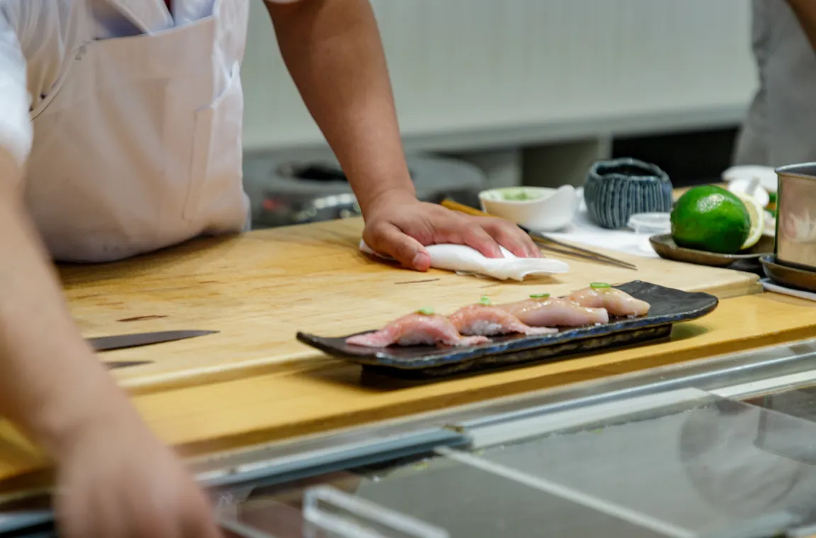 A sushi chef wiping down a light wooden counter next to a long black plate featuring four pieces of freshly prepared nigiri sushi.