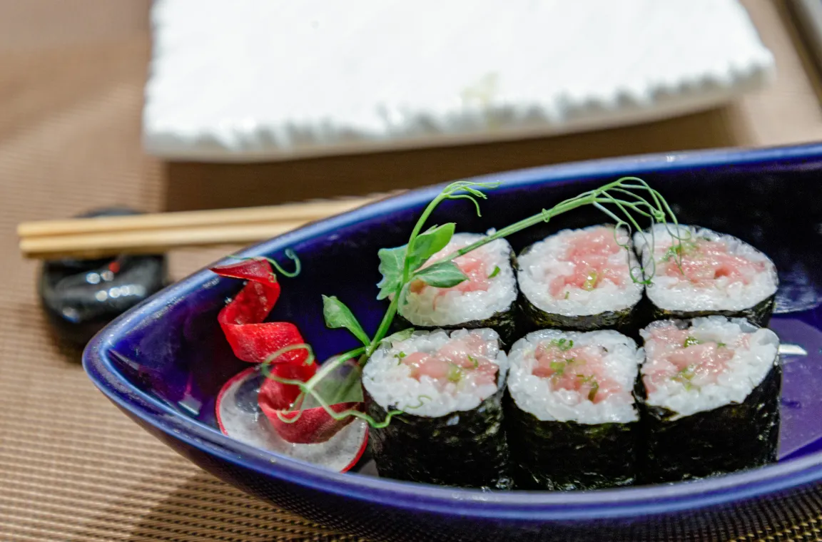 Six pieces of tuna maki rolls neatly arranged in a blue curved ceramic dish, garnished with a sprig of greenery and a curled beet ribbon next to chopsticks.