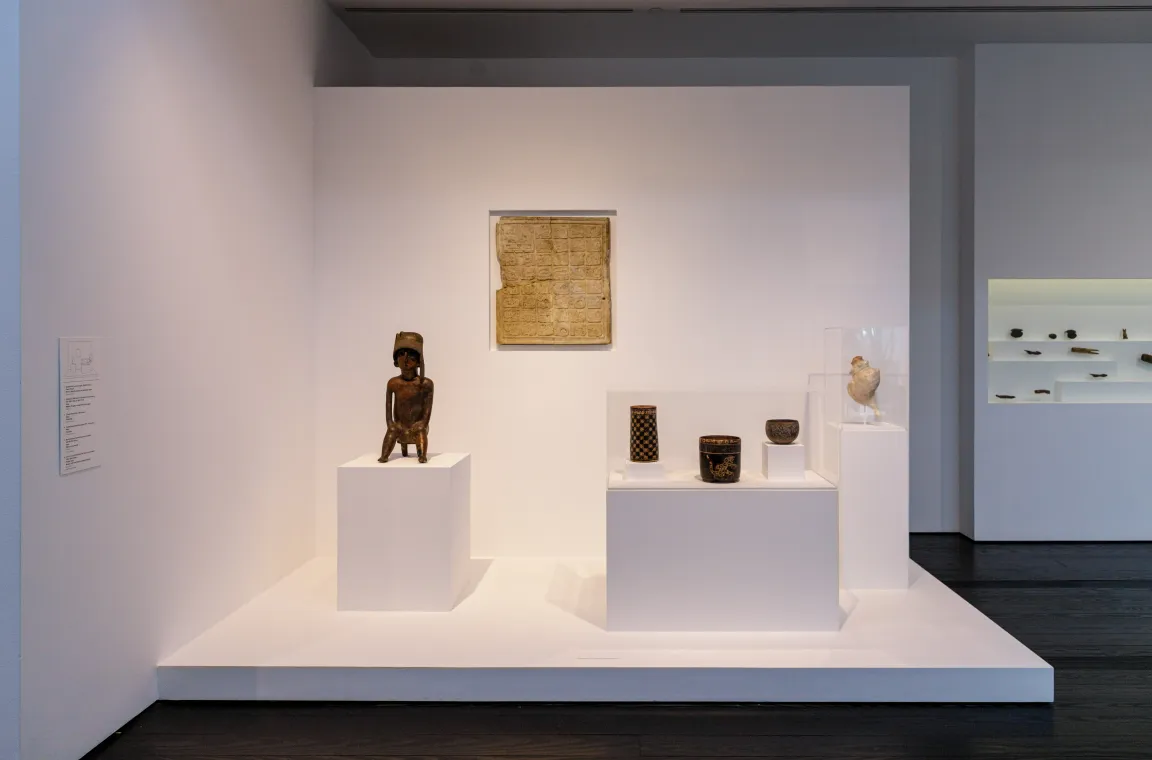 An indoor museum exhibit displaying ancient artifacts on white pedestals, including a small dark sculpted figure, small decorative cups, and a stone tablet mounted on the wall.