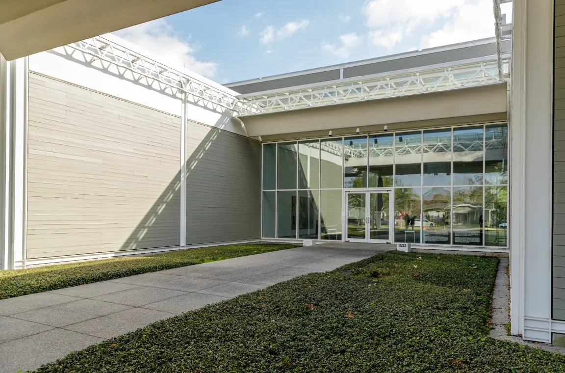 Exterior view of a modern pavilion entrance at The Menil Collection, featuring gray horizontal siding, a large glass window facade, a concrete walkway, and manicured green groundcover.