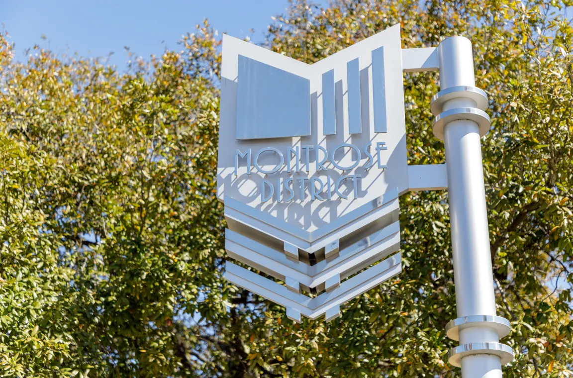 A shiny metallic street sign that reads "MONTROSE DISTRICT" mounted on a silver pole, set against a backdrop of lush green trees.