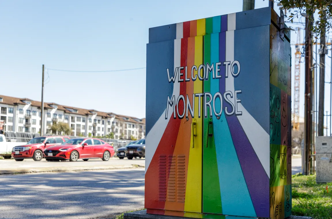 A metal utility box painted with a vibrant rainbow sunburst pattern and the words "WELCOME TO MONTROSE," with a street and parked cars in the background.